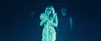 Movie still from “Jem and the Holograms” (2015), directed by Jon M. Chu – A woman in a white dress holding a microphone in front of two men; Wide shot, Over the shoulder angle