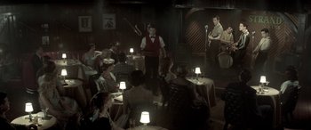 Movie still from “Jersey Boys” (2014), directed by Clint Eastwood – A group of people sitting at a table in front of a microphone; Wide shot, Over the shoulder angle