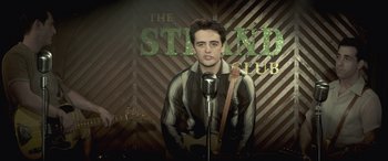 Movie still from “Jersey Boys” (2014), directed by Clint Eastwood – A man holding a guitar and a microphone in front of a microphone stand; Medium shot, Low angle