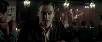 Movie still from “Jersey Boys” (2014), directed by Clint Eastwood – A man in a suit and tie sitting in front of a chandelier; Close Up shot, Over the shoulder angle