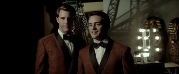 Movie still from “Jersey Boys” (2014), directed by Clint Eastwood – Two men in suits posing for a picture in front of a bridge; Medium shot, Low angle
