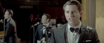 Movie still from “Jersey Boys” (2014), directed by Clint Eastwood – A man in a suit and tie holding a microphone; Close Up shot, Over the shoulder angle