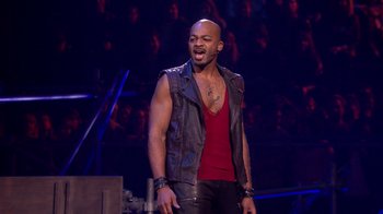Movie still from “Jesus Christ Superstar Live in Concert” (2018), directed by Alex Rudzinski – A man in a red shirt and black leather vest singing; Medium shot, Overhead angle