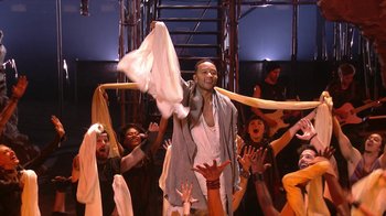 Movie still from “Jesus Christ Superstar Live in Concert” (2018), directed by Alex Rudzinski – A group of people are gathered around a man holding up a towel; Wide shot, Low angle