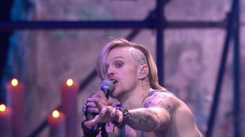 Movie still from “Jesus Christ Superstar Live in Concert” (2018), directed by Alex Rudzinski – A man with long blonde hair is singing into a microphone; Medium shot, Over the shoulder angle