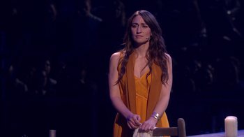 Movie still from “Jesus Christ Superstar Live in Concert” (2018), directed by Alex Rudzinski – A woman in an orange dress standing in front of an audience; Medium shot, Over the shoulder angle