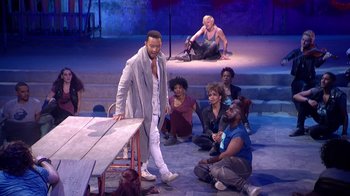 Movie still from “Jesus Christ Superstar Live in Concert” (2018), directed by Alex Rudzinski – A group of people sitting on the ground in front of a table; Wide shot, High angle