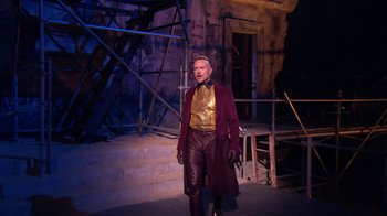 Movie still from “Jesus Christ Superstar Live in Concert” (2018), directed by Alex Rudzinski – A man wearing a red coat and a gold shirt standing in front of a building; Wide shot, Low angle