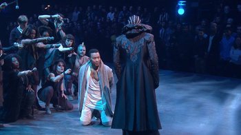 Movie still from “Jesus Christ Superstar Live in Concert” (2018), directed by Alex Rudzinski – A man kneeling down in front of another man in front of an audience; Wide shot, High angle