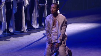 Movie still from “Jesus Christ Superstar Live in Concert” (2018), directed by Alex Rudzinski – A man standing on a stage holding a cane; Medium shot, High angle