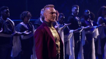 Movie still from “Jesus Christ Superstar Live in Concert” (2018), directed by Alex Rudzinski – A man in a red jacket standing in front of a group of people; Medium shot, Over the shoulder angle