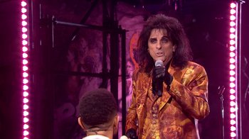 Movie still from “Jesus Christ Superstar Live in Concert” (2018), directed by Alex Rudzinski – A man in a yellow and red suit holding a microphone; Medium shot, Over the shoulder angle