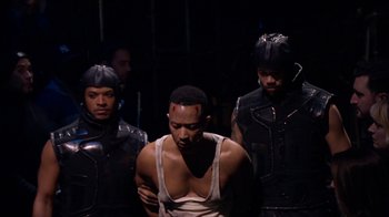 Movie still from “Jesus Christ Superstar Live in Concert” (2018), directed by Alex Rudzinski – A group of men standing next to each other in a dark room; Medium shot, Over the shoulder angle