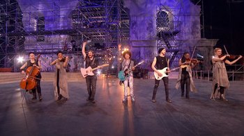 Movie still from “Jesus Christ Superstar Live in Concert” (2018), directed by Alex Rudzinski – A group of people standing on a stage with guitars; Wide shot, Low angle