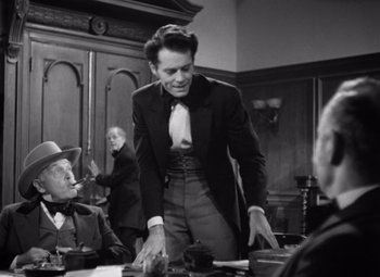 Movie still from “Jezebel” (1938), directed by William Wyler – A man in a suit and tie standing at a table with other men; Medium shot, Over the shoulder angle