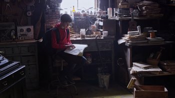 Movie still from “Turkey Hollow” (2015), directed by Kirk R. Thatcher – A person sitting at a desk in a room with lots of clutter; Wide shot, Low angle