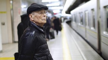 Movie still from “Jiro Dreams of Sushi” (2011), directed by David Gelb – An older man wearing glasses and a hat waiting for a train; Close Up shot, Low angle