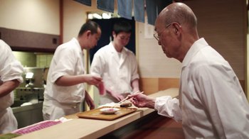 Movie still from “Jiro Dreams of Sushi” (2011), directed by David Gelb – A man in a white shirt is cutting food with two other men; Medium shot, Over the shoulder angle