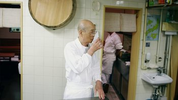 Movie still from “Jiro Dreams of Sushi” (2011), directed by David Gelb – An older man in a white uniform drinking a cup of coffee; Medium shot, Over the shoulder angle
