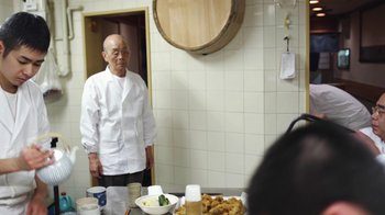 Movie still from “Jiro Dreams of Sushi” (2011), directed by David Gelb – An older man standing in front of a table of food; Medium shot, Over the shoulder angle