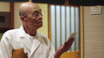 Movie still from “Jiro Dreams of Sushi” (2011), directed by David Gelb – An older man holding a cell phone in his hands; Medium shot, Low angle