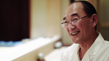 Movie still from “Jiro Dreams of Sushi” (2011), directed by David Gelb – An older man wearing glasses sitting in a room; Close Up shot, Low angle