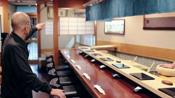 Movie still from “Jiro Dreams of Sushi” (2011), directed by David Gelb – A person sitting at a long table in front of a row of chairs; Medium shot, Over the shoulder angle
