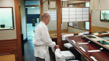Movie still from “Jiro Dreams of Sushi” (2011), directed by David Gelb – A man standing in front of a table in a restaurant; Medium shot, Over the shoulder angle