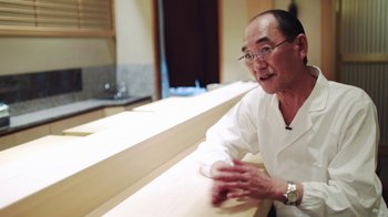 Movie still from “Jiro Dreams of Sushi” (2011), directed by David Gelb – A man in glasses and a white shirt is talking; Medium shot, High angle