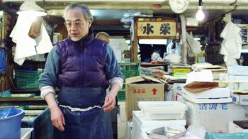 Movie still from “Jiro Dreams of Sushi” (2011), directed by David Gelb – A man standing in front of a pile of food; Medium shot, Low angle