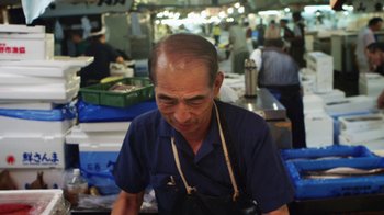 Movie still from “Jiro Dreams of Sushi” (2011), directed by David Gelb – An older man in an apron is in a fish market; Medium shot, High angle