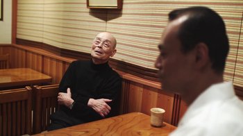 Movie still from “Jiro Dreams of Sushi” (2011), directed by David Gelb – A man sitting at a table talking to another man in front of him; Medium shot, Over the shoulder angle