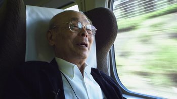 Movie still from “Jiro Dreams of Sushi” (2011), directed by David Gelb – An older man sitting in a chair on a train; Medium shot, Low angle