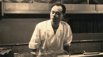 Movie still from “Jiro Dreams of Sushi” (2011), directed by David Gelb – A man wearing a white shirt is standing in front of a window; Medium shot, Low angle