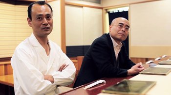 Movie still from “Jiro Dreams of Sushi” (2011), directed by David Gelb – Two men sitting at a table in front of a mirror; Medium shot, Low angle