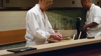Movie still from “Jiro Dreams of Sushi” (2011), directed by David Gelb – An old man in a white shirt and white jacket is working in a kitchen; Medium shot, Low angle