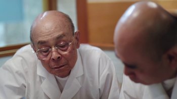 Movie still from “Jiro Dreams of Sushi” (2011), directed by David Gelb – An older man in a white lab coat looking at himself in the mirror; Close Up shot, Low angle