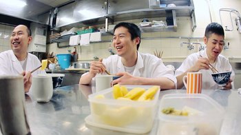 Movie still from “Jiro Dreams of Sushi” (2011), directed by David Gelb – A man sitting at a table in a kitchen with food; Medium shot, Low angle