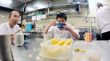 Movie still from “Jiro Dreams of Sushi” (2011), directed by David Gelb – A man sitting at a table in a kitchen holding a bowl; Medium shot, Low angle