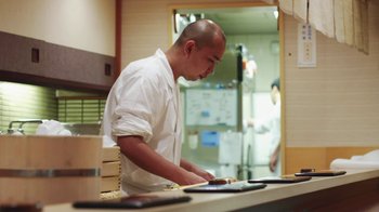 Movie still from “Jiro Dreams of Sushi” (2011), directed by David Gelb – A man standing in front of a counter preparing food; Medium shot, Low angle