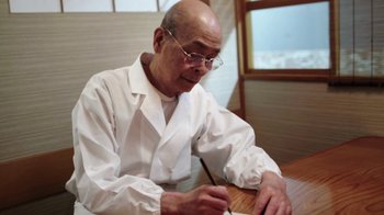 Movie still from “Jiro Dreams of Sushi” (2011), directed by David Gelb – An older man sitting at a table writing on a piece of paper; Medium shot, Low angle