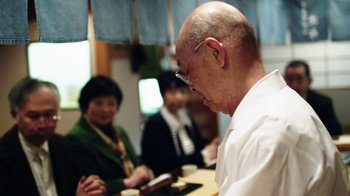 Movie still from “Jiro Dreams of Sushi” (2011), directed by David Gelb – An older man with glasses is looking down at a table; Medium shot, Over the shoulder angle
