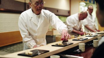 Movie still from “Jiro Dreams of Sushi” (2011), directed by David Gelb – A man in a white lab coat is cutting food; Wide shot, High angle