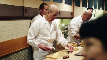 Movie still from “Jiro Dreams of Sushi” (2011), directed by David Gelb – An older man in a white shirt and white apron cutting food in a kitchen; Medium shot, Low angle