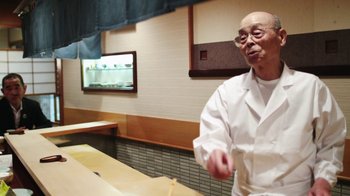 Movie still from “Jiro Dreams of Sushi” (2011), directed by David Gelb – An older man in a white lab coat is holding chopsticks in his hand; Medium shot, Low angle