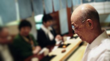 Movie still from “Jiro Dreams of Sushi” (2011), directed by David Gelb – An old man is sitting at a table with other people; Extreme Close Up shot, Over the shoulder angle