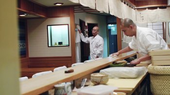 Movie still from “Jiro Dreams of Sushi” (2011), directed by David Gelb – A man standing in front of a counter with other people; Wide shot, Low angle