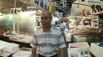 Movie still from “Jiro Dreams of Sushi” (2011), directed by David Gelb – A man standing in an asian market with fish on the ground; Medium shot, Low angle