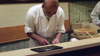 Movie still from “Jiro Dreams of Sushi” (2011), directed by David Gelb – A man in a white lab coat is cutting food on a tablet computer; Medium shot, Low angle