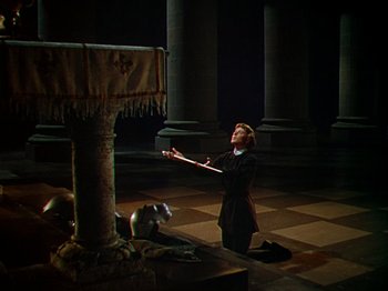 Movie still from “Joan of Arc” (1948), directed by Victor Fleming – A woman kneeling on the ground in a dark room; Wide shot, High angle